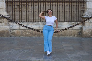 Kot pantolon, beyaz tişört ve güneş gözlüğü giymiş güzel genç bir kadın Seville 'deki gotik katedrali çevreleyen zincirlerde oturuyor. Kadın şehre yaptığı ziyaretten zevk alıyor. Turizm konsepti