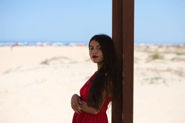 Genç ve güzel, uzun saçlı Latin bir kadın sahildeki ahşap bir afişe yaslanıyor. Arka planda şemsiyeli kum tepeleri deniz ve ufukta mavi gökyüzü.