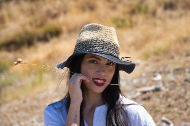 Beyaz gömlek ve hasır şapka giymiş, güzel, genç, esmer bir kadının portresi. Arka planda ağaçlar ve bitki örtüsü var.