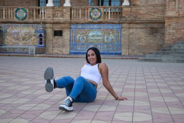 Genç ve güzel Fas 'lı kadın beyaz bluz ve kot pantolon giymiş Sevilla' da bir ayağı havada yerde oturuyor. Kadın İspanyol şehrinde tatilin tadını çıkarıyor.