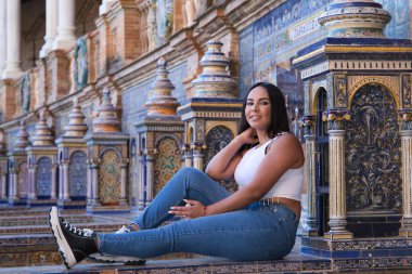 Genç, güzel, esmer Fas 'lı kadın beyaz bluz ve kot pantolon giymiş Seville' de İspanya meydanında fayanslı bir bankta oturuyor. Kadın mutlu ve gülümsüyor. Seyahat ve turizm kavramı