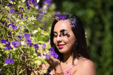 Genç, güzel, koyu renk saçlı kadın bir demet menekşe çiçeği alıyor, onlara bakıyor ve onları kokluyor, kadın rahatlamış, mutlu ve gülümsüyor. Mutluluğu ve sarı günü kavra