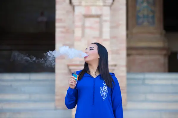 Faslı bir kadın, genç ve güzel, mavi abaya giyiyor, tipik Arap kostümü, elektrikli sigarayla sigara içiyor. 31 Mayıs, Dünya Tütünsüz Günü. Akciğer hastalıkları ve kanser kavramı