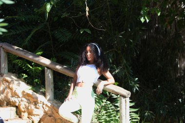 Genç, güzel, Latin, kıvırcık saçlı, spor kıyafetleri giymiş, ahşap çitlere yaslanarak beyaz kulaklıklarla müzik dinliyor.