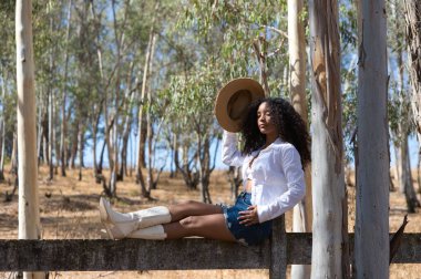 Kadın, genç, güzel, Latin, kıvırcık saçlı, beyaz gömlekli, kot etekli, beyaz çizmeli ve şapkalı, bacakları okaliptüs ormanının çitlerinde uzanmış oturuyor.