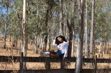 Kadın, genç, güzel, Latin, kıvırcık saçlı, beyaz gömlekli, kot etekli, beyaz çizmeli ve şapkalı, bacaklarını orman çitlerine uzatmış oturuyor. Kadın dinleniyor.