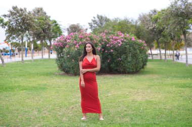 Uzun saçlı, yeşil gözlü, çok zarif kırmızı elbiseli genç ve güzel İspanyol kadın parkta çimlerin üzerinde manken gibi yürüyor ve farklı vücut duruşları sergiliyor.