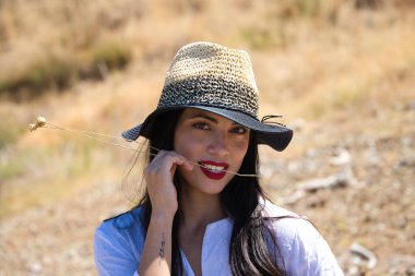 Beyaz gömlek ve hasır şapka giymiş, güzel, genç, esmer bir kadının portresi. Arka planda ağaçlar ve bitki örtüsü var.