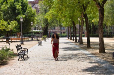 Büyük kahverengi gözlü, kıvırcık saçlı, kırmızı elbiseli güzel Latin bir kadın parktaki ağaçların arasında yürüyor ve manken gibi farklı pozlar veriyor. Moda ve güzellik konsepti