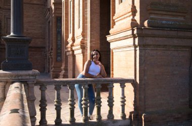 Beyaz bluz ve kot pantolon giymiş genç ve güzel Faslı esmer kadın Sevilla 'daki İspanya meydanının taş korkuluklarında yüzünü dinlendiriyor. Kadın mutlu ve gülümsüyor.