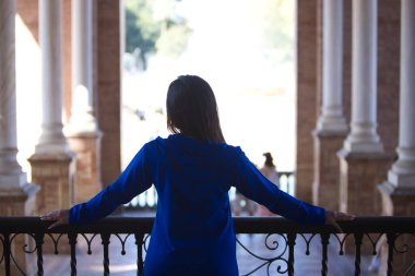 Faslı bir kadın, esmer, abaya 'da giyinmiş genç ve güzel Seville' deki meydanın balkonuna yaslanmış kolonlar ve kemerler arasından karşıya bakıyor. Fotoğraf arkadan çekilmiş.