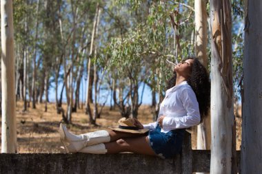 Kadın, genç, güzel, Latin, kıvırcık saçlı, beyaz gömlekli, kot etekli, beyaz çizmeli ve şapkalı, bacaklarını orman çitlerine uzatmış oturuyor. Kadın gözlerini kapar.