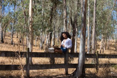 Kadın, genç, güzel, Latin, kıvırcık saçlı, beyaz gömlekli, kot etekli, beyaz çizmeli ve şapkalı, bacaklarını orman çitlerine uzatmış oturuyor. Kadın dinleniyor.