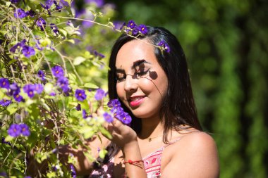 Genç, güzel, koyu renk saçlı kadın bir demet menekşe çiçeği alıyor, onlara bakıyor ve onları kokluyor, kadın rahatlamış, mutlu ve gülümsüyor. Mutluluğu ve sarı günü kavra