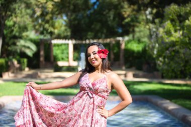 Genç, güzel esmer kadın, uzun çiçekli elbisesi ve saçında çiçek deseni var. Kadın elbisesini eliyle kaldırır ve gülümser. Tatil ve seyahat kavramı