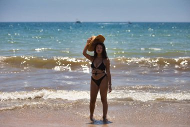Kahverengi gözlü genç ve güzel esmer kadın sahilde mayo giyiyor ve kafasına hasır bir şapka takıyor ve elleriyle tutuyor. Arka planda deniz ve mavi gökyüzü olan ufuk.