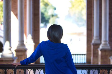 Faslı bir kadın, esmer, abaya 'da giyinmiş genç ve güzel Seville' deki meydanın balkonuna yaslanmış kolonlar ve kemerler arasından karşıya bakıyor. Fotoğraf arkadan çekilmiş.