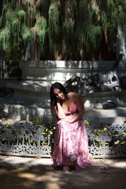Genç, güzel, koyu saçlı bir kadın bahçenin kenarında oturuyor bacakları çapraz ve yanakları elinde dinleniyor. Kadın düşünüyor ve yüzünde ciddi bir ifade var.