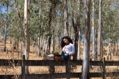 Kadın, genç, güzel, Latin, kıvırcık saçlı, beyaz gömlekli, kot etekli, beyaz çizmeli ve şapkalı, bacaklarını orman çitlerine uzatmış oturuyor. Kadın dinleniyor.