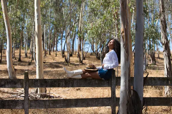 Kadın, genç, güzel, Latin, kıvırcık saçlı, beyaz gömlekli, kot etekli, beyaz çizmeli ve şapkalı, bacaklarını orman çitlerine uzatmış oturuyor. Kadın gözlerini kapar.