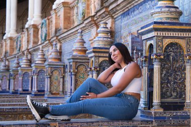 Genç, güzel, esmer Fas 'lı kadın beyaz bluz ve kot pantolon giymiş Seville' de İspanya meydanında fayanslı bir bankta oturuyor. Kadın mutlu ve gülümsüyor. Seyahat ve turizm kavramı
