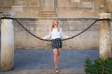 Genç ve güzel sarışın bir kadın, beyaz bir gömlek ve beyaz benekli kısa siyah bir etek giyiyor, Sevilla katedralini çevreleyen büyük demir bir zincir üzerinde ellerini tutuyor ve yaslanıyor.