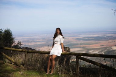 Güzel esmer Latin kadın, İspanya 'da Endülüs' ün doğal bir bakış açısıyla ahşap bir korkulukta oturuyor. Kadın gülümser, beyaz ve altın ayakkabılar giyer.