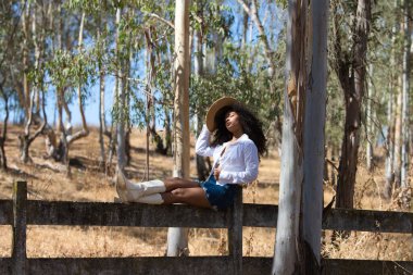 Kadın, genç, güzel, Latin, kıvırcık saçlı, beyaz gömlekli, kot etekli, beyaz çizmeli ve şapkalı, bacakları okaliptüs ormanının çitlerinde uzanmış oturuyor.