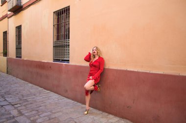Sarışın bir kadın, genç ve güzel, makyaj, kırmızı ve zarif giyinmiş, kırmızı ve pembe bir duvara yaslanmış, bir bacağını duvara dayayıp farklı jestler ve vücut ifadeleri yapıyor.