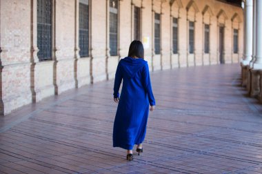 Mavi abaya giymiş Faslı esmer kadın, tipik Arap kostümü Seville 'deki İspanyol meydanında kemer ve sütunlar arasında yürüyor. Fotoğraf arkadan çekilmiş. Seyahat ve tatil konsepti