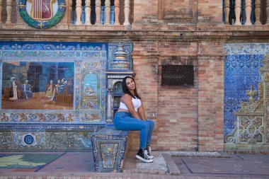 Genç, güzel, esmer Fas 'lı kadın beyaz bluz ve kot pantolon giymiş Seville' de İspanya meydanında fayanslı bir bankta oturuyor. Kadın mutlu ve gülümsüyor. Seyahat ve turizm kavramı