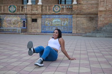 Genç ve güzel Fas 'lı kadın beyaz bluz ve kot pantolon giymiş Sevilla' da bir ayağı havada yerde oturuyor. Kadın İspanyol şehrinde tatilin tadını çıkarıyor.