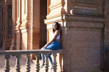 Genç, güzel, siyah saçlı, beyaz bluzlu ve kot pantolonlu kadın Sevilla 'daki İspanyol meydanının taş korkuluklarında oturuyor. Kadın mutlu ve gülümsüyor. Seyahat ve turizm kavramı