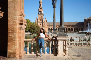 Beyaz tişörtlü, Faslı genç ve güzel bir kadın ve İspanyol meydanındaki korkuluklardan birine yaslanmış kot pantolonlar, kulelerinden birinin ve kanalın arka planında Seville 'de. Seyahat kavramı