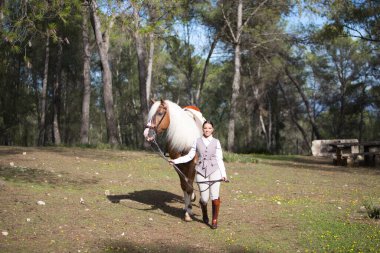Genç ve güzel bir binici kadın dizginleri elinde tutuyor ve safkan kahverengi atıyla bir çam ormanında yürüyor. Kadın çok mutlu. Ata duyulan sevgi kavramı