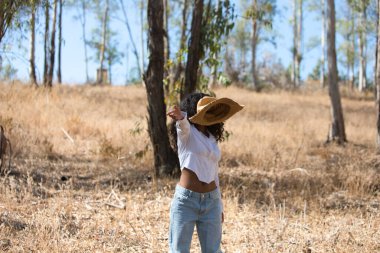 Kadın, genç, güzel, Latin, kıvırcık saçlı, beyaz gömlekli, kot pantolonlu ve botlu esmer, uçan bir disk gibi dönen hasır bir şapka fırlatıyor. Çiftçi okaliptüs ağaçlarının arasında.