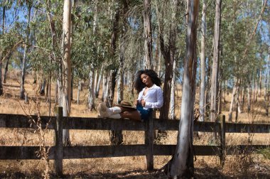 Kadın, genç, güzel, Latin, kıvırcık saçlı, beyaz gömlekli, kot etekli, beyaz çizmeli ve şapkalı, bacaklarını orman çitlerine uzatmış oturuyor. Kadın dinleniyor.