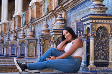 Genç, güzel, esmer Fas 'lı kadın beyaz bluz ve kot pantolon giymiş Seville' de İspanya meydanında fayanslı bir bankta oturuyor. Kadın mutlu ve gülümsüyor. Seyahat ve turizm kavramı