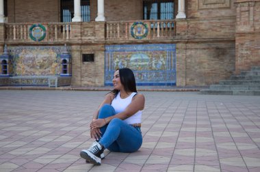 Faslı esmer kadın, genç ve güzel, beyaz bluzlu ve kot pantolonlu, yerde oturuyor ve kollarıyla bacağını tutuyor, kız Seville 'in kare İspanya' sı. Seyahat ve turizm kavramı