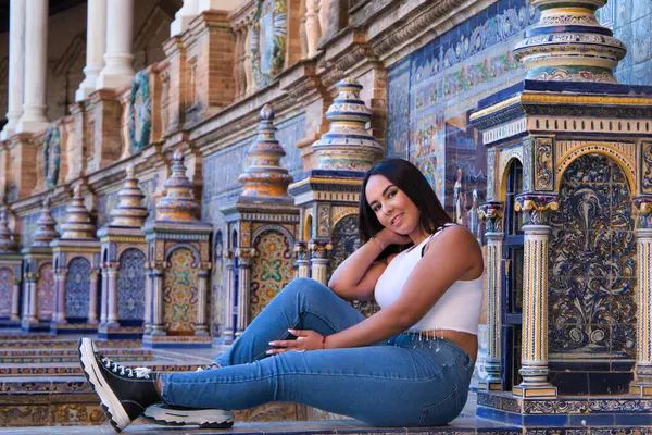 Genç, güzel, esmer Fas 'lı kadın beyaz bluz ve kot pantolon giymiş Seville' de İspanya meydanında fayanslı bir bankta oturuyor. Kadın mutlu ve gülümsüyor. Seyahat ve turizm kavramı