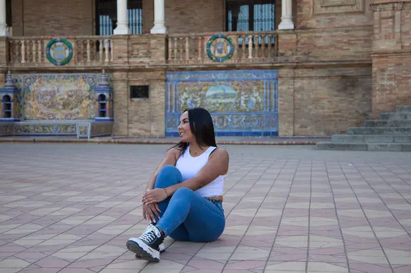 Faslı esmer kadın, genç ve güzel, beyaz bluzlu ve kot pantolonlu, yerde oturuyor ve kollarıyla bacağını tutuyor, kız Seville 'in kare İspanya' sı. Seyahat ve turizm kavramı