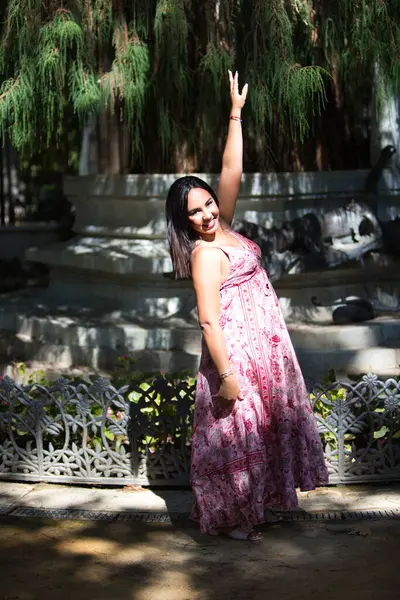 Çiçek desenli uzun elbiseli güzel, esmer bir kadın. Elbisesini elinde tutuyor ve bir kolunu kaldırıyor. Arka planda bataklık selvisi, taksonyum distichum. Kadın mutlu.