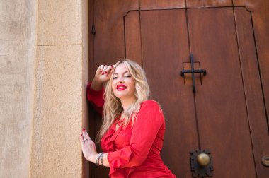 Sarışın, genç ve güzel, makyajlı, kırmızı ve zarif, kollarını duvara dayamış ve kameraya şehvetli ve mutlu bir şekilde bakıyor. Güzellik ve moda konsepti