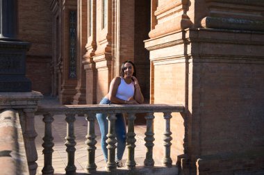 Beyaz bluz ve kot pantolon giymiş genç ve güzel Faslı esmer kadın Sevilla 'daki İspanya meydanının taş korkuluklarında yüzünü dinlendiriyor. Kadın mutlu ve gülümsüyor.