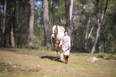 Genç ve güzel bir binici kadın dizginleri elinde tutuyor ve safkan kahverengi atıyla bir çam ormanında yürüyor. Kadın çok mutlu. Ata duyulan sevgi kavramı