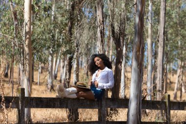 Kadın, genç, güzel, Latin, kıvırcık saçlı, beyaz gömlekli, kot etekli, beyaz çizmeli ve şapkalı, bacaklarını orman çitlerine uzatmış oturuyor. Kadın dinleniyor.