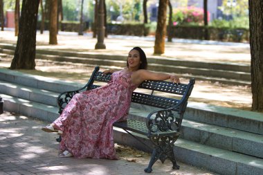 Genç, güzel esmer bir kadın parkta bir bankta oturuyor, kolları sırtüstü uzanıyor, bacakları çapraz ve başı arkaya eğik.