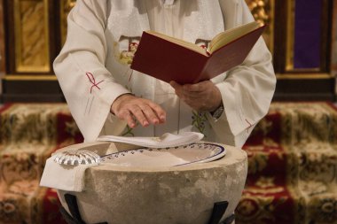 The priest blesses with his hands the water of the baptismal font to proceed to the sacrament of baptism in a catholic church. On the edge of the font the silver shell and the white handkerchief clipart