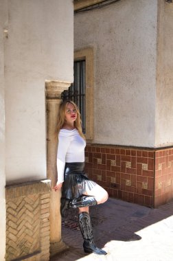 Beyaz tişörtlü, deri etekli, siyah çizmeli güzel sarışın bir kadın bir bacağını taştan bir sütuna koyar. Kadın ciddi.