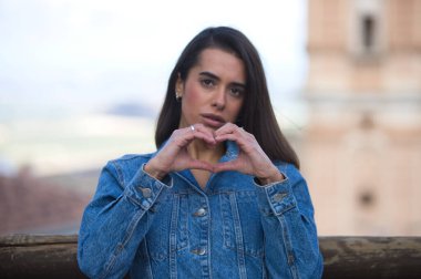 Latin kadın, genç ve güzel kot gömlek giymiş, parmakları ve elleri ile kalp yapıyor ve gülümsüyor.